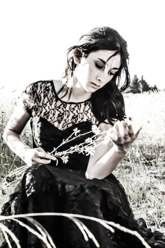 Photo of a young woman in a black laced dress, looking down at flowers she is holding with a compassionate concerned look on her face