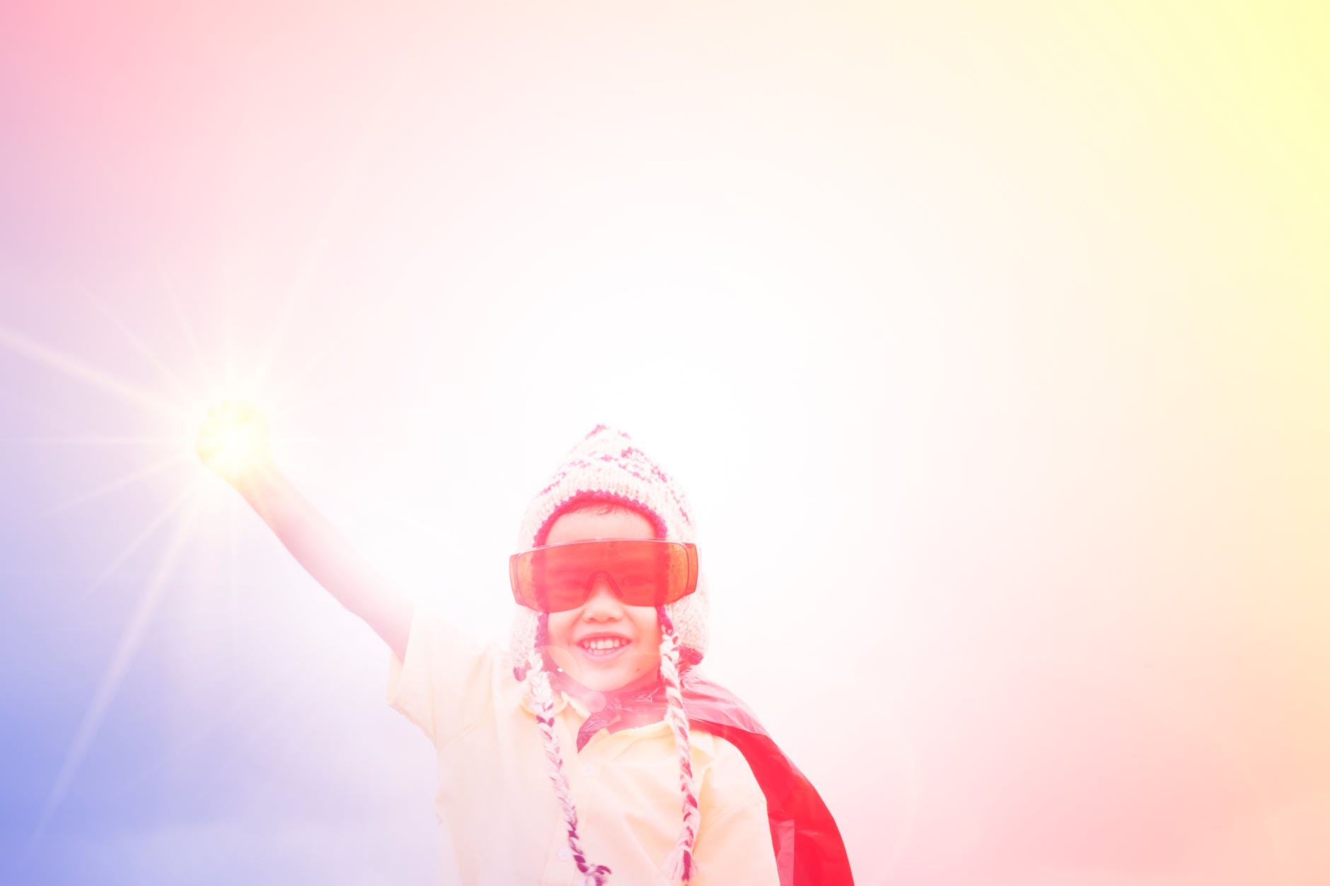 Photo of a boy with his hand in the air like super man, wearing a cape and sunglasses that are too big for his face, but with a really big smile on his face.